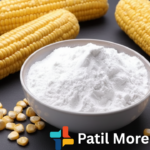 A bowl of white cornstarch sits on a dark tabletop, surrounded by several ears of fresh yellow corn and loose corn kernels.